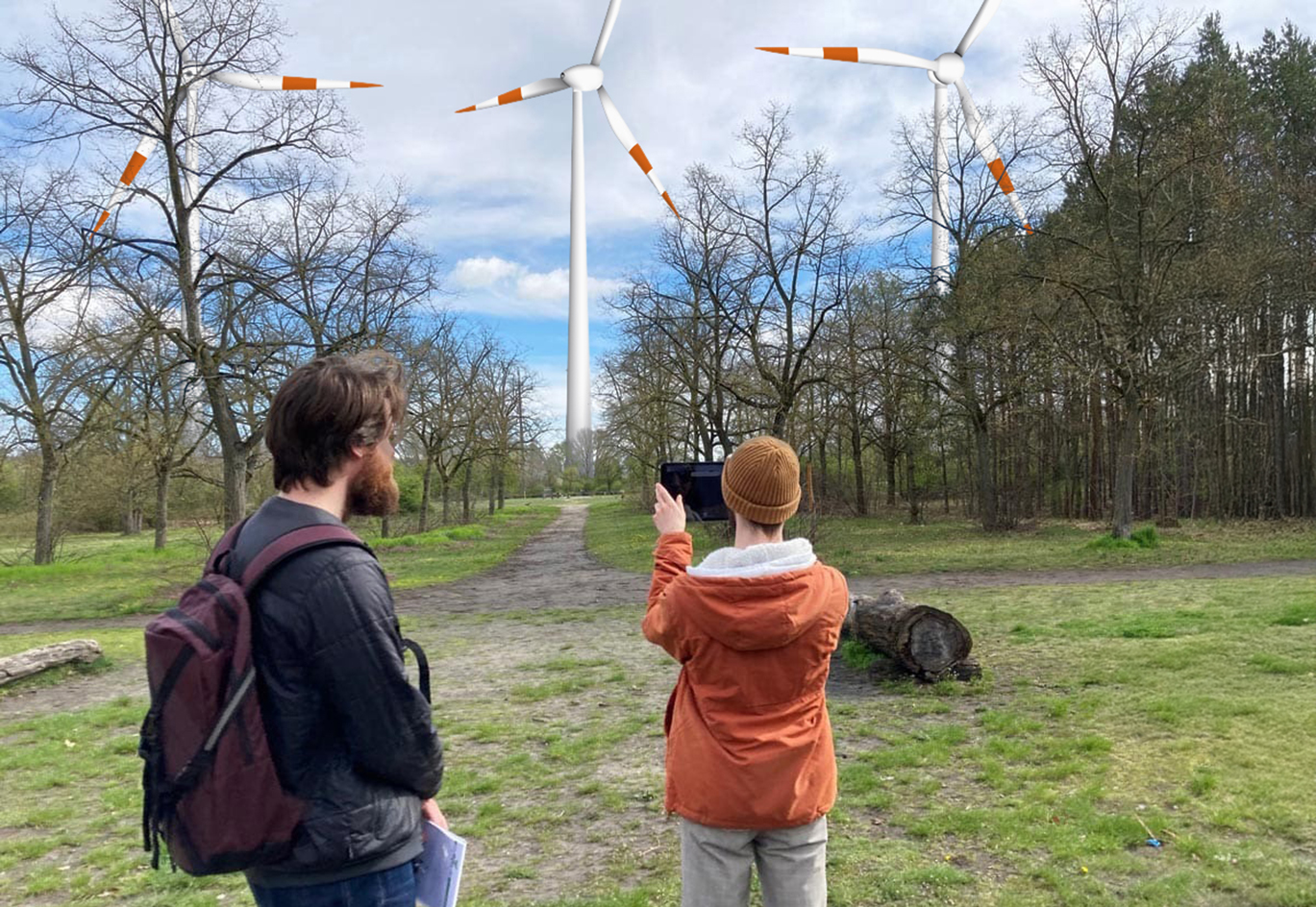 Männer mit Tablet auf einer Wiese fotografieren Windräder in einiger Entfernung. Visualisiert das Projekt AR4WIND.