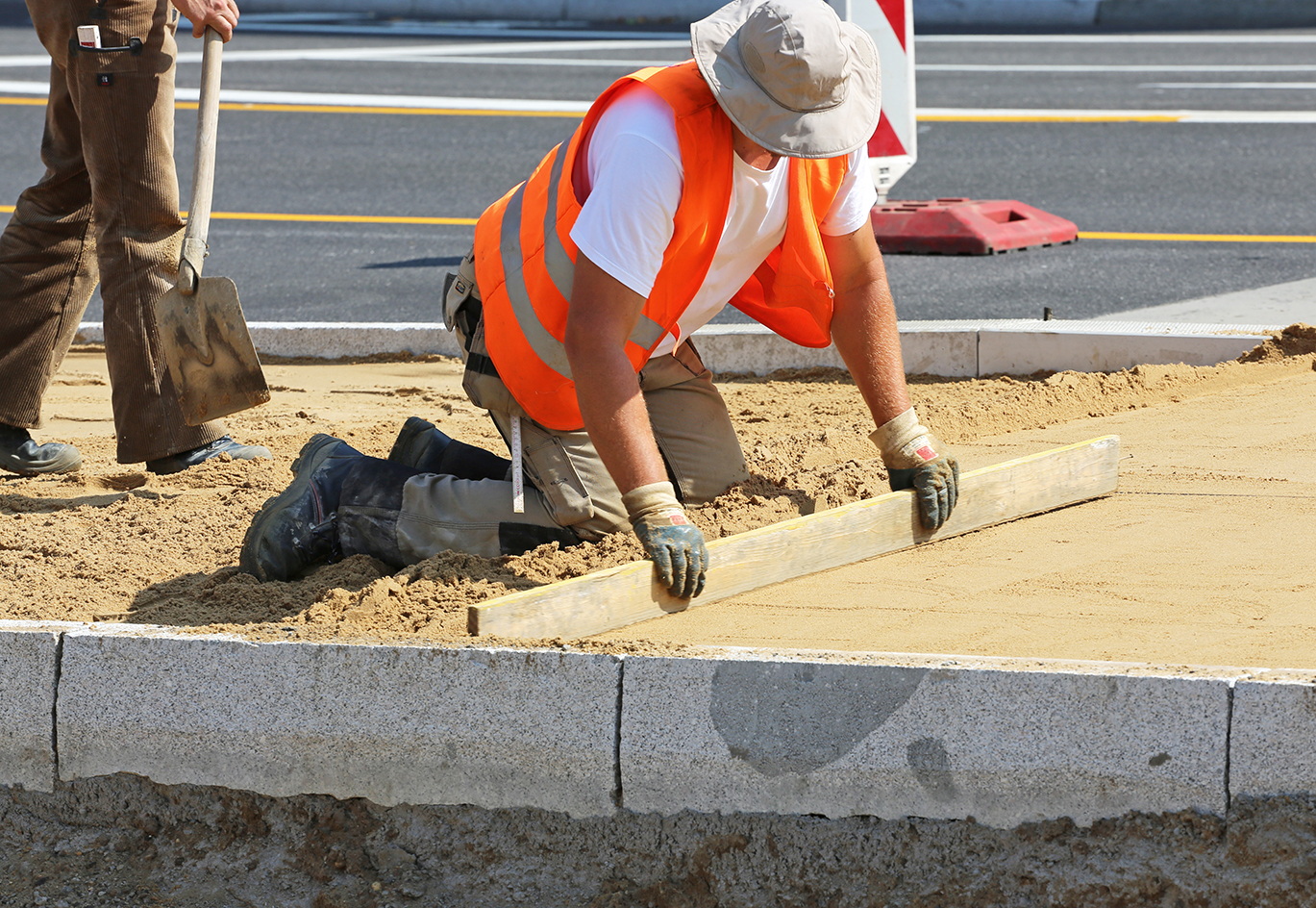 Ein Straßenarbeiter zieht bei Hitze das Pflasterbett auf einer Baustelle ab. Zum Schutz trägt er einen Sonnenhut. Visualisiert das Projekt Adapt2Care.