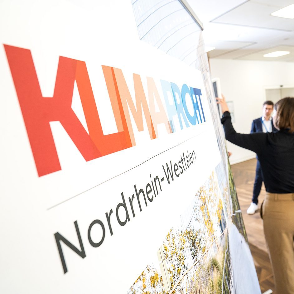 Frau präsentiert vor einer Gruppe von Menschen an einer Stellwand mit dem Projekttitel. Visualisiert das Projekt KLIMA.PROFIT NRW